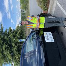 Jennifer Helgeson, holding a clipboard and wearing a high-vis vest stands next to a car. The car has a large sign on the side that reads “Center for Community Resilience Planning. Lumberton, NC Flood Study Team.” 