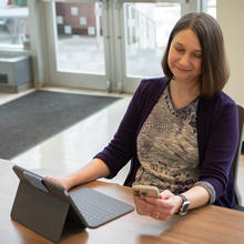 Julie Haney sits at a table in the NIST library with a laptop, looking at a smartphone in her hand. 