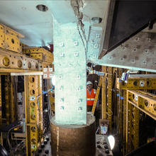 Heavy equipment and scaffolding surrounds a vertical concrete slab and attached horizontal beam, which is being cracked away.