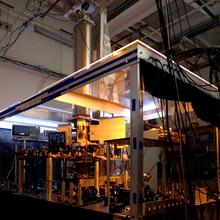 A large clear-sided box inside a darkened lab holds electronic equipment, wiring, and a metal cylinder rising up from the box. 