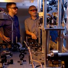Two researchers in safety glasses stand behind a table holding various electronic and laser devices. A metal structure in the foreground is part of the atomic clock. 