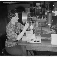 Historical photo shows woman adjusting a large metal lamp at a table full of similar equipment. 