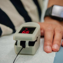 Close-up of a clip-on medical device on the finger of a person sitting on an exam table. The device screen reads 99. 