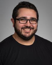 headshot of man with dark hair and beard wearing black shirt and glasses