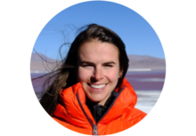 person with long brown hair wearing a bright orange puffy coat standing in front of a mountain landscape