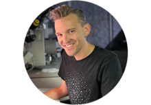Andrew D. Iams, a person with short dirty blonde hair, sitting at a computer and smiling