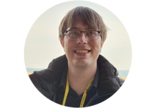 Benjamin Dolata, a person with short brown hair and glasses, standing outdoors and smiling