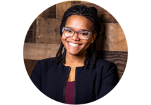 Headshot of a woman with long black braided hair, wearing, glasses, dressed professionally, and smiling for the photo.