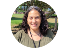 person with long curly black hair and safety glasses on her head, smiling in front of a tree