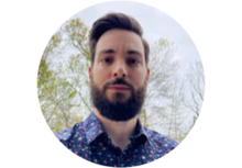 person with dark brown hair and a beard, wearing a dress shirt and standing in front of trees