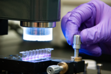 A hand in a purple glove turns a metal dial on a plate holding tiny vials of purple liquid. 