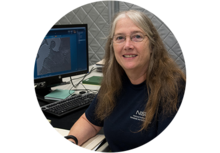 NIST's Maureen Williams, a person with long brown/gray hair and glasses, sits at a computer with technical graphics and smiles for the camera