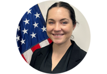 Headshot of Carolyn Pace, a woman with black hair, pinned up, wearing a black cardigan, standing in front of an American flag