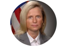 Headshot of Dianne Poster, woman with long blonde hair, wearing a white shirt and gray suit jacket, in front of an American flag