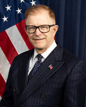 Headshot of a man wearing glasses and a dark suit with an American flag behind him