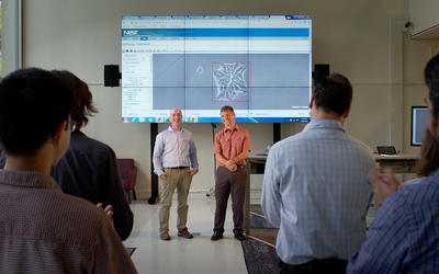Two men stand in front of an audience, demonstrating WIPP, which is projected via computer onto several large screens in a library.