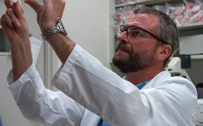 A man in a lab coat and safety glasses looks up at petri dishes he is holding. 