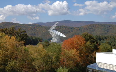 A white telescope dish is surrounded by wooded mountains in autumn colors.