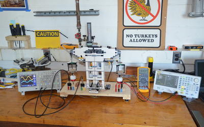 A Lego structure wired to various electronic readout devices stands on a wooden counter surrounded by other scientific equipment. A sign on the concrete wall behind reads "No Turkeys Allowed."