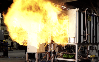 A fireball rushes out of the opening of a small metal compartment in a concrete room. 