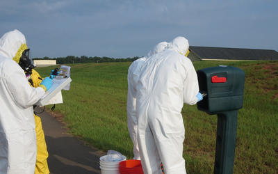 Four figures in coveralls stand outdoors around a mailbox, looking at a clipboard and other items. 