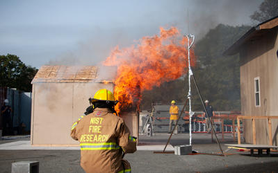 Photo shows a NIST firefighter on standby watching a shed burn next to a target structure representing a wall of a residence during an experiment.