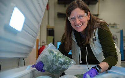 Melissa Phillips, wearing safety glasses, poses leaning over an open chest freezer, holding a plastic bag of spinach. 