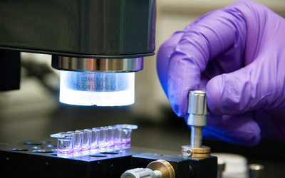 A hand in a purple glove turns a metal dial on a plate holding tiny vials of purple liquid. 