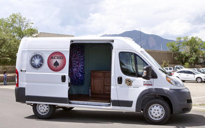 A white van with its sliding door open, parked in a lot, with mountains in the background. Decals on the side refer to scientific studies.