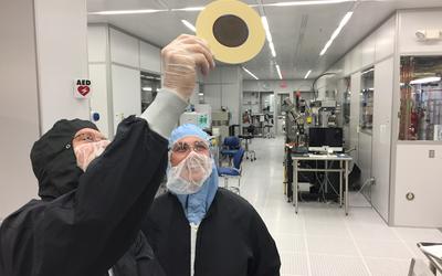 Two people wearing coveralls in a "clean room" lab look up at a disk-shaped object that one of them is holding. 