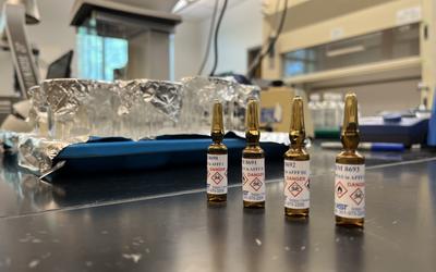 Four small dark glass ampules stand on a lab table, market with skull and crossbones danger icons. 