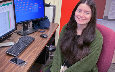 Isabelle Rivera poses smiling at a computer with two monitors, one showing a spreadsheet and the other multiple file icons. 