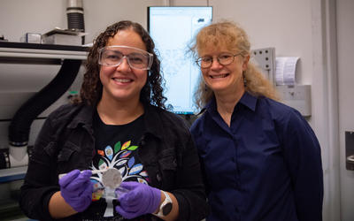 Diana Ortiz-Montalvo, wearing safety glasses and holding a small circular filter with tweezers, poses with Abigail Lindstrom in the lab.