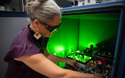A researcher wearing safety glasses reaches into a box of circuitry and other equipment, which emits a green glow. 