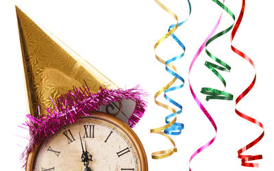An antique clock showing a few minutes before 12 is topped with a party hat, and colorful streamers hang in the background. 