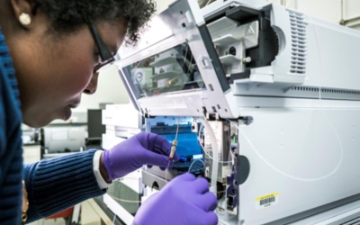 A researcher leans close to an open boxlike device, handling small tubes and vials inside.