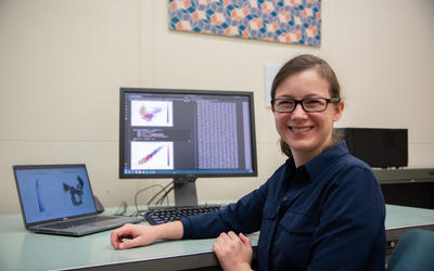 Melinda Kleczynski poses sitting at her desk, with a monitor showing data and graphs behind her. 