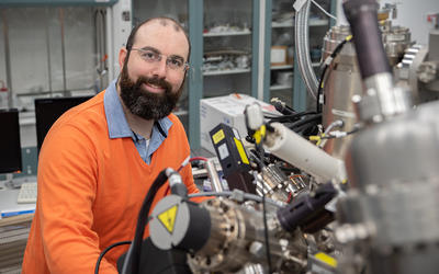 Trey Diulus poses sitting in the lab with a large, complex scientific instrument in the foreground.