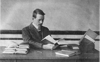 Historical photo shows Henry Moseley sitting at a desk reading a thick book, with other open books on the desk. 