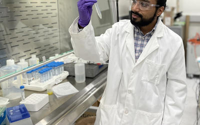 George Caceres wears safety glasses and a lab coat as he looks at a plastic test tube he is holding.