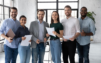 Six smiling, casually dressed young people stand in a line in an office space, holding papers and laptops.