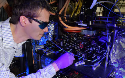 Stephen Eckel wears dark safety glasses as he peers into the complex CAVS apparatus in a darkened lab. 