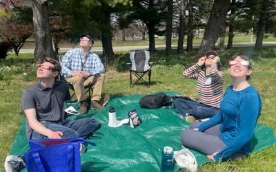 Four people sitting outside on a green tarp wear eclipse glasses as they peer up into the sky. 