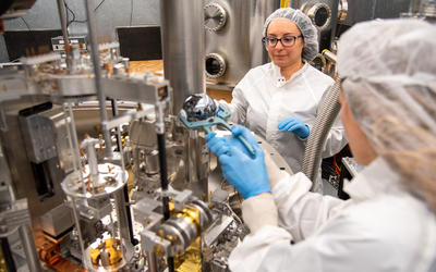 A researcher wearing a lab coat uses tongs to place a silvery sphere into a large scientific device as another researcher looks on. 
