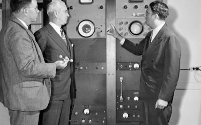 Historical photo shows three men standing next to a refrigerator-sized piece of machinery with an analog clock attached to the top. 