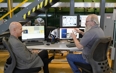 Two men sit at a table in a lab, facing each other and smiling as they talk and gesture toward their computer screens. 