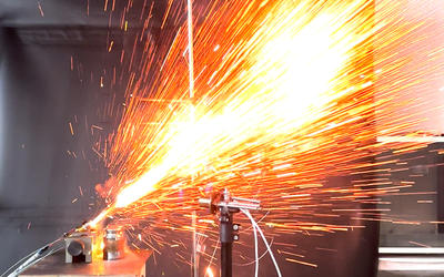 An explosion of sparks shoots upward at a 45 degree angle from a AA lithium battery clamped into place. The explosion takes place in a laboratory surrounded by safety and measurement equipment.