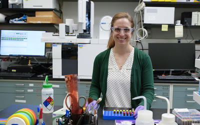 Briana Capistran wears safety glasses as she poses smiling in the lab with computers and other equipment in the background. 