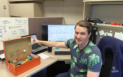 Sam Schaffter poses sitting in his office with a shoebox modified with craft supplies to be an educational model of an E. coli cell. 
