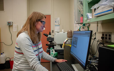 A scientist wearing safety glasses stands in front of a monitor in her lab.
